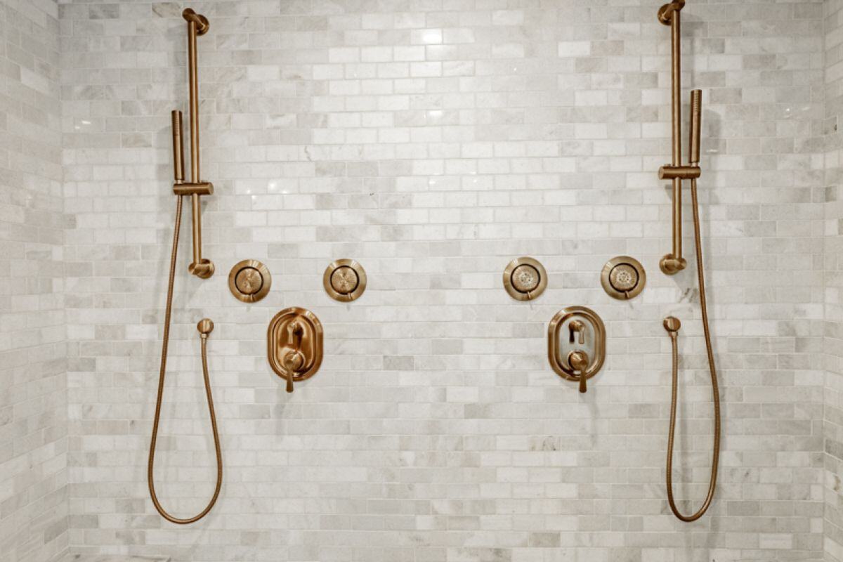 A luxury shower setup featuring two brass handheld showerheads and elegant control knobs mounted on marble tiles. | Ever After Homes