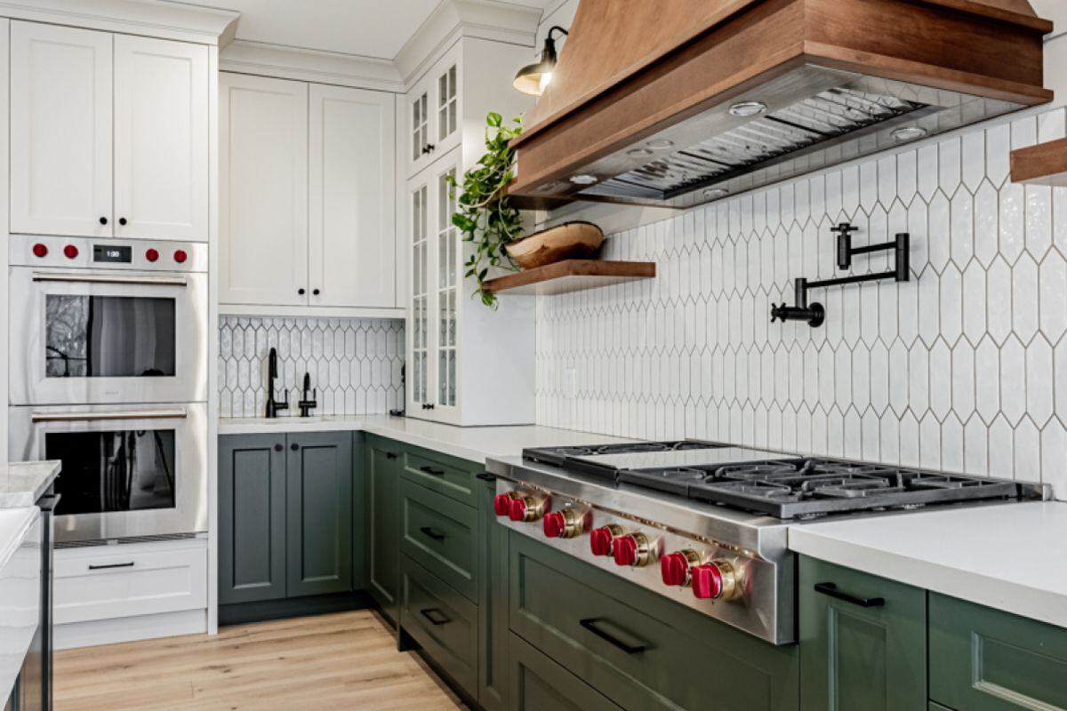 A sleek kitchen featuring green lower cabinets, white upper cabinets, a wooden hood vent, and a hexagonal tile backsplash. | Ever After Homes