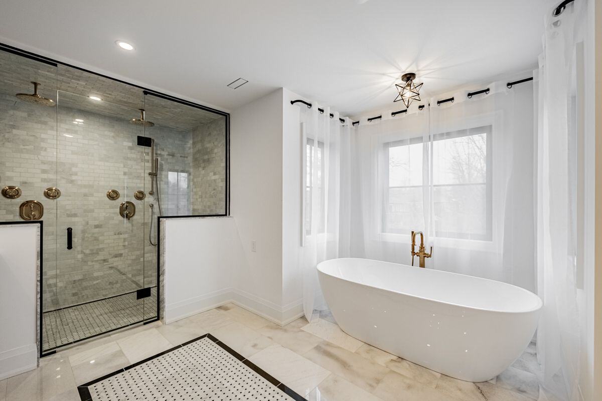 A bright bathroom with a freestanding white tub, brass faucet, and a walk-in shower with marble tile and glass enclosure. | Ever After Homes