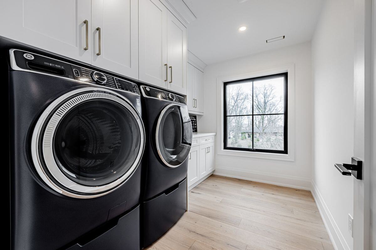 A modern laundry room with black front-loading washer and dryer, white cabinetry, and a large black-framed window. | Ever After Homes