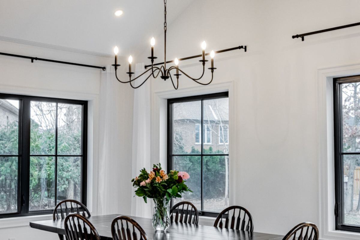 A close-up of a dining room with a simple black chandelier, a dark wood table, and large windows providing outdoor views. | Ever After Homes