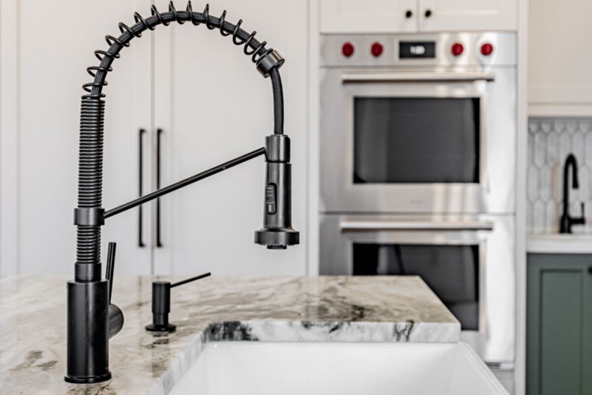 A close-up of a black kitchen faucet with a spring design, set against a marble countertop and stainless steel double ovens in the background. | Ever After Homes