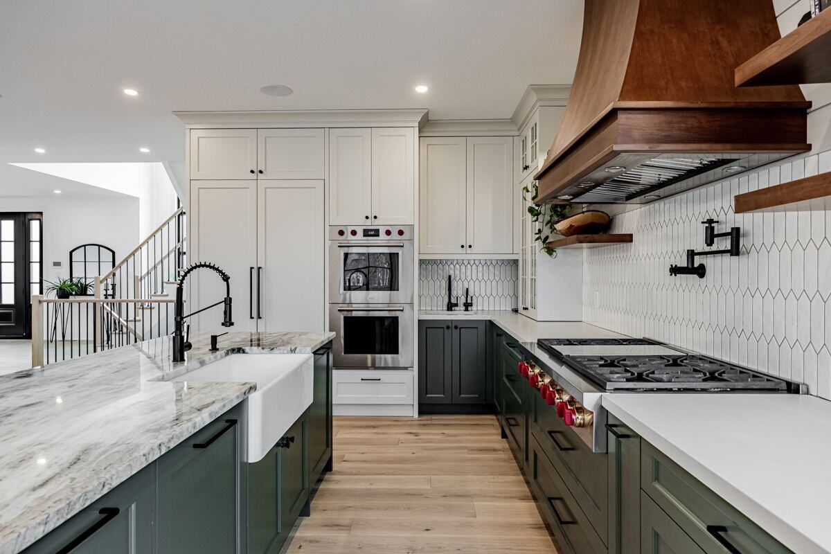 A modern kitchen with white cabinets, dark green lower cabinetry, a marble countertop, and a farmhouse sink. The space includes a stainless steel double oven and a large wooden hood vent. | Ever After Homes