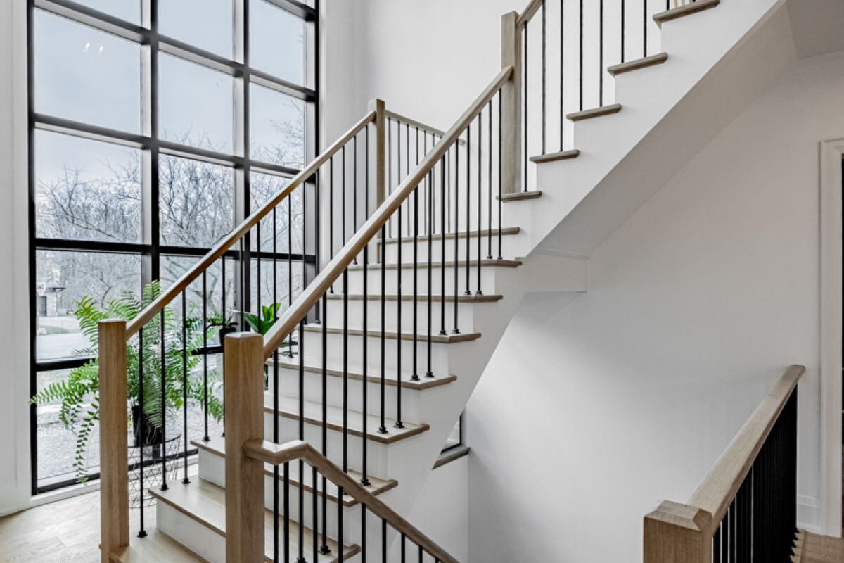 A detailed view of a staircase with black iron railings and large windows overlooking a scenic outdoor area. The natural wood accents provide a warm touch to the modern design. | Ever After Homes