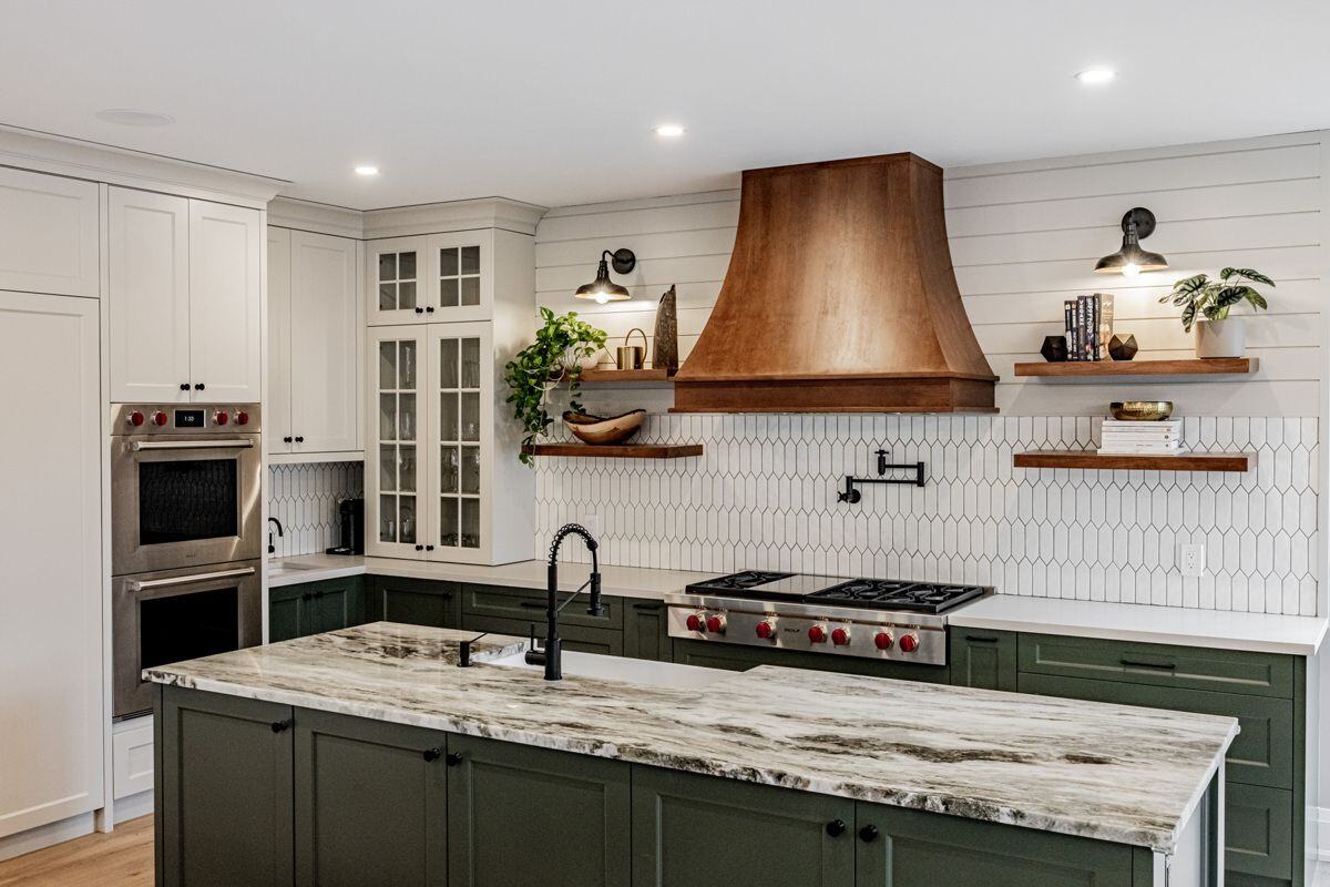 A kitchen with a mix of white and green cabinetry, open wooden shelving, and a large wooden hood vent. The marble island counter and modern black fixtures add a touch of elegance. | Ever After Homes