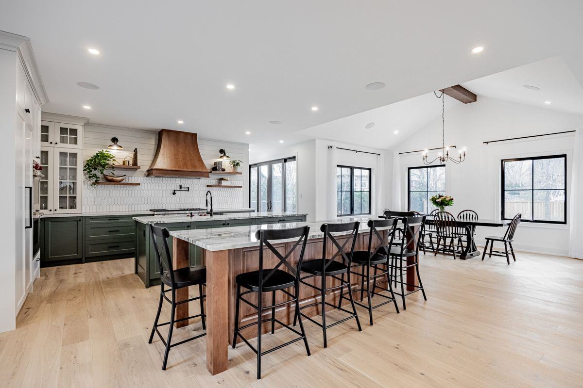 An open-concept kitchen and dining area with a large marble island, black barstools, and a dining table surrounded by windows. The space feels bright and inviting with natural light. | Ever After Homes