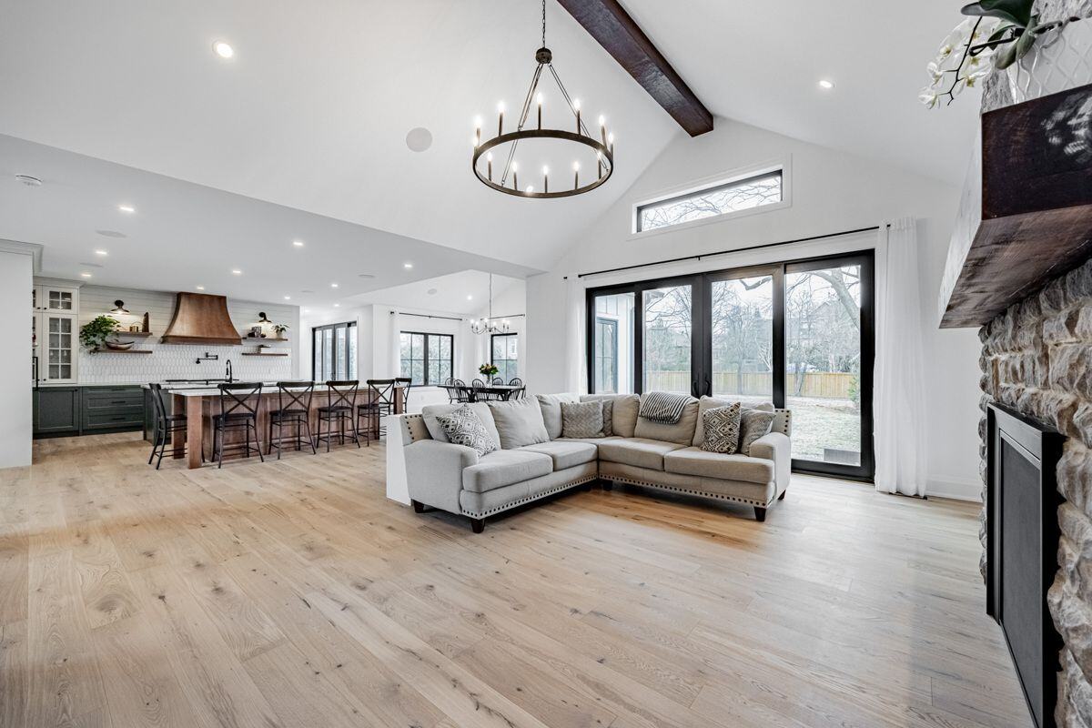 A spacious living room with a stone fireplace, a wooden mantle, and a cozy sectional sofa. Large windows bring in plenty of natural light, complementing the neutral-toned decor. | Ever After Homes