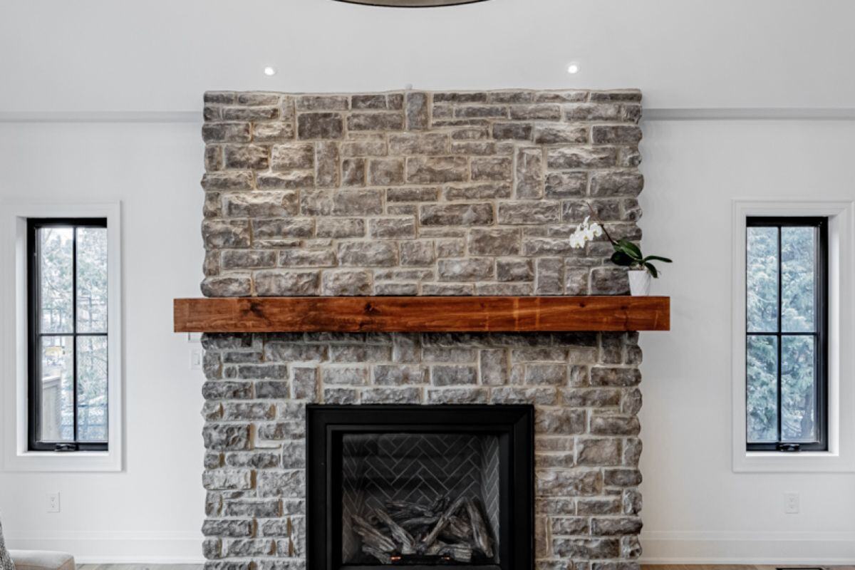 A close-up of a stone fireplace with a wooden mantle, featuring a small orchid and modern black-framed windows on either side. The textured stone adds a rustic charm. | Ever After Homes