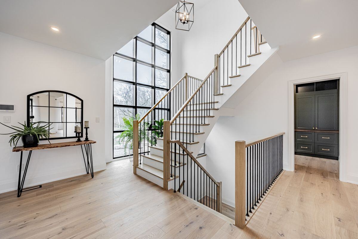 A staircase with wooden steps, black metal railings, and a large floor-to-ceiling grid-style window that lets in ample natural light. A small console table with plants and decor completes the space. | Ever After Homes